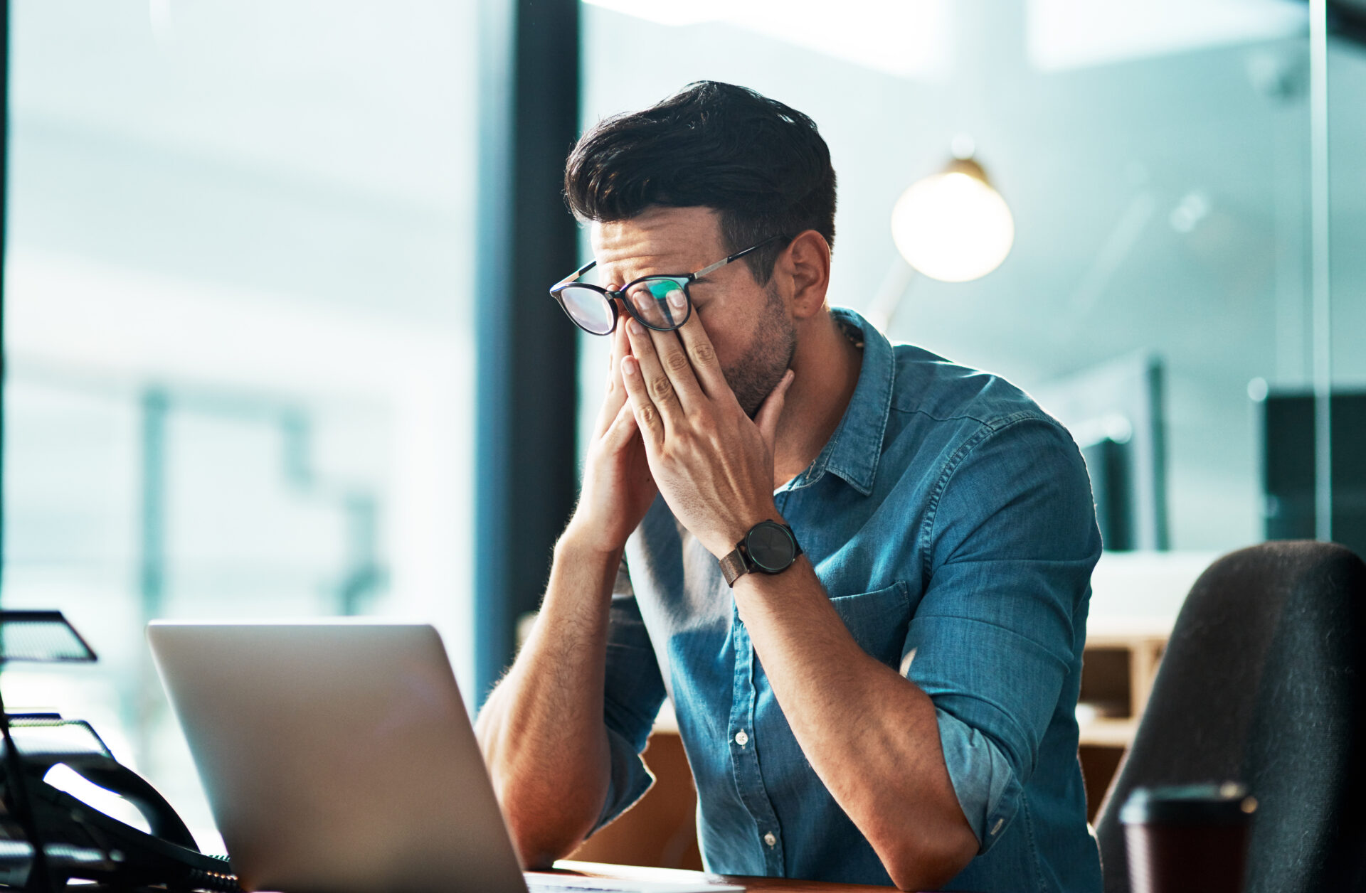 A man in glasses and a denim shirt sits at a desk, rubbing his eyes in stress before an open laptop, with blurred office lights behind.