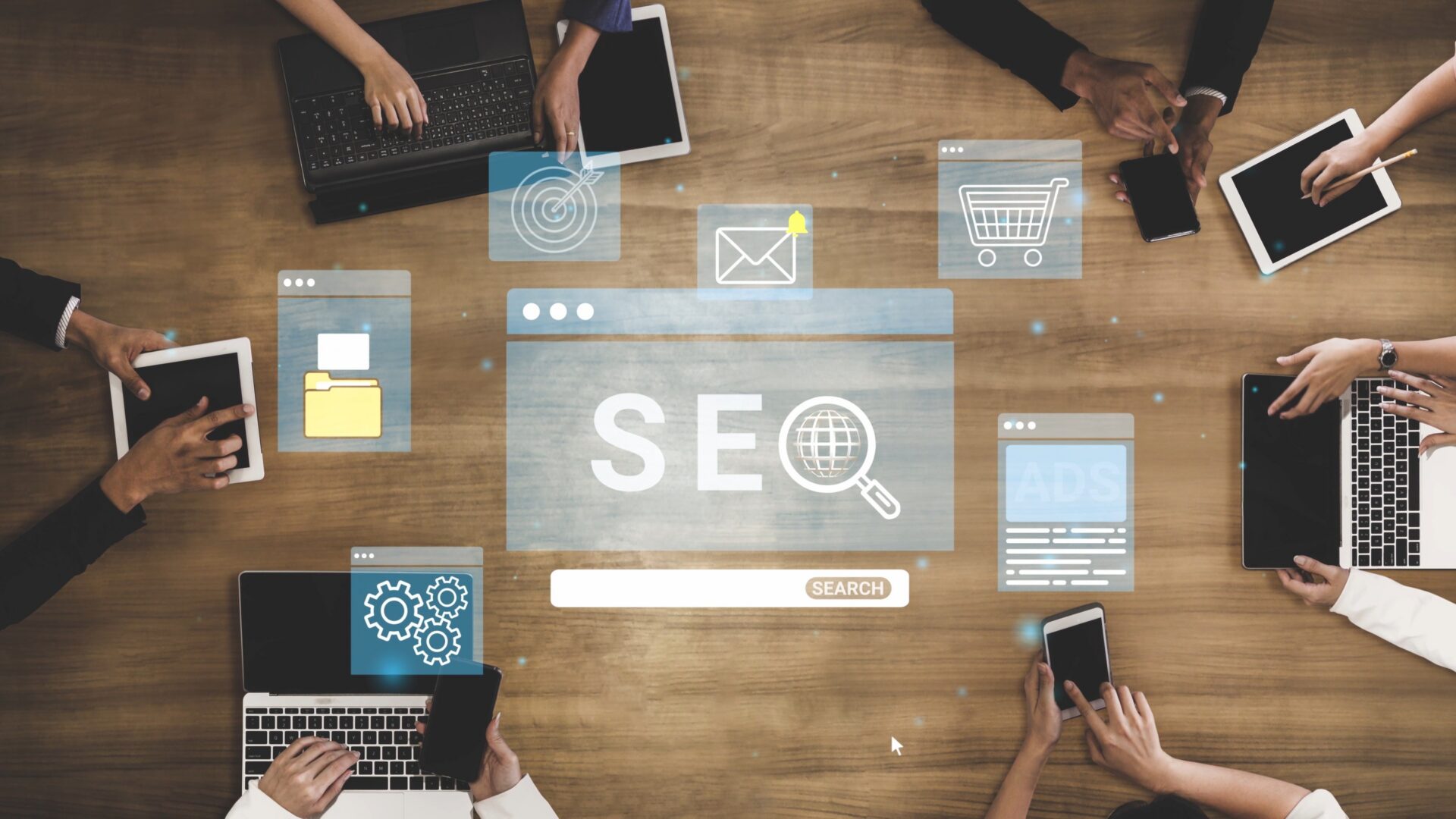 Top-down view of people at a table using devices, with digital icons for SEO, email, shopping cart, gears, and ads around them. Indicates SEO strategy.
