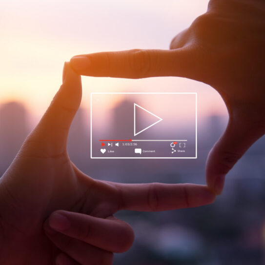 Two hands shape a rectangle framing a blurred sunset cityscape, with a digital play button and video controls centered over the image. Indicates a video marketing strategy.