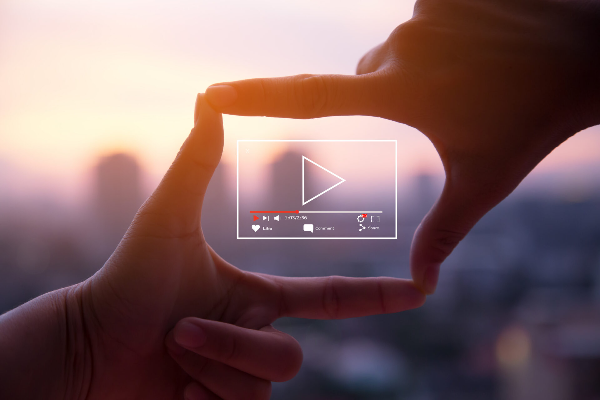Two hands shape a rectangle framing a blurred sunset cityscape, with a digital play button and video controls centered over the image. Indicates a video marketing strategy.