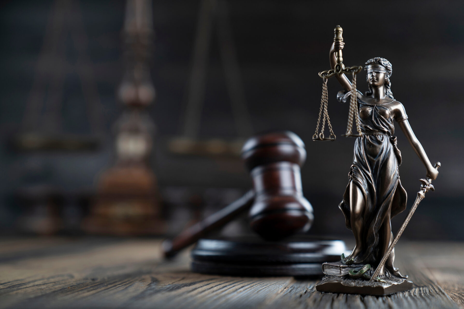 Bronze Lady Justice statue with scales and sword stands on desk near judge’s gavel, blurred legal symbols seen in the background.