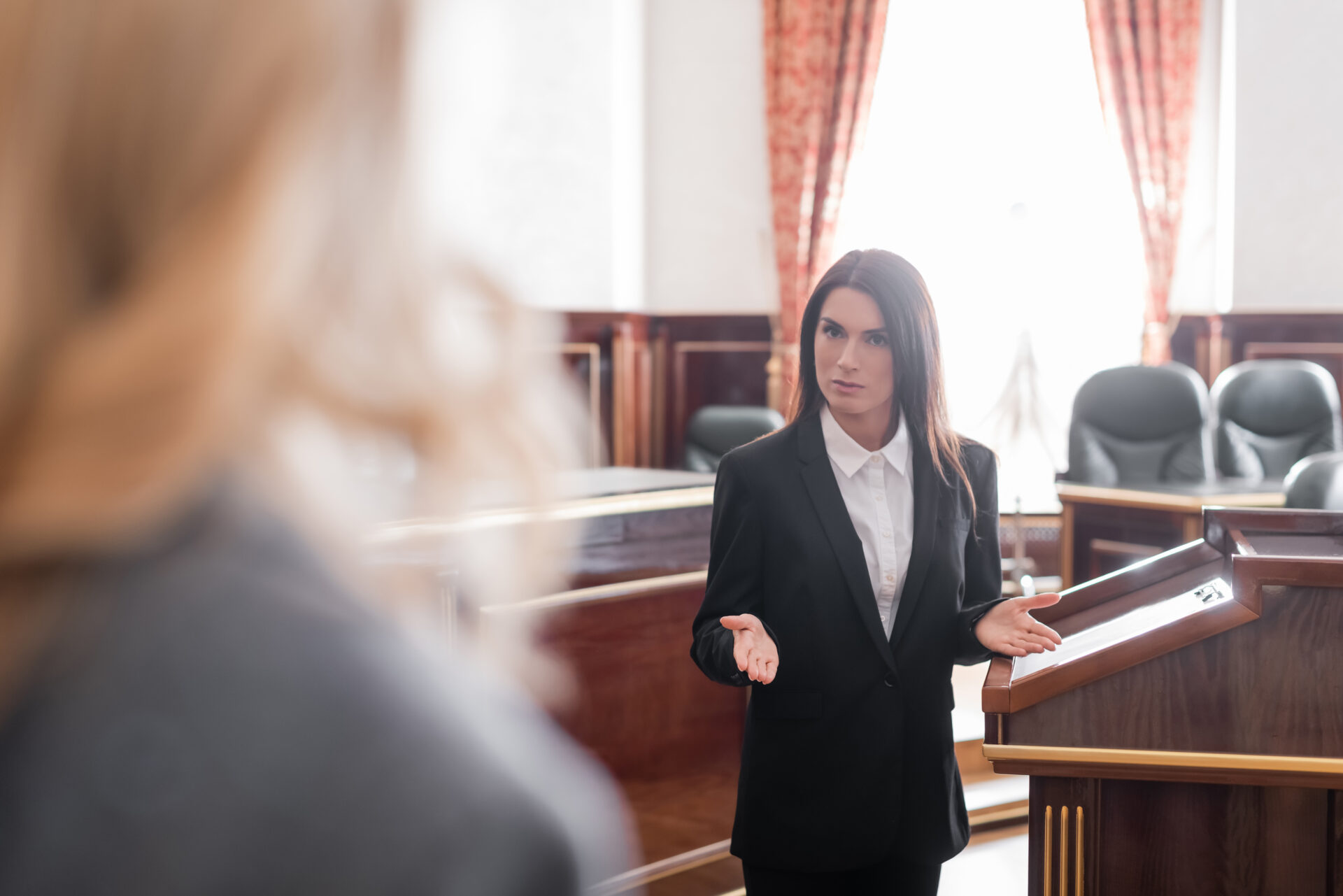 A woman in a black suit stands at a courtroom lectern, gesturing as she addresses someone whose back is seen. Empty seats and windows behind.