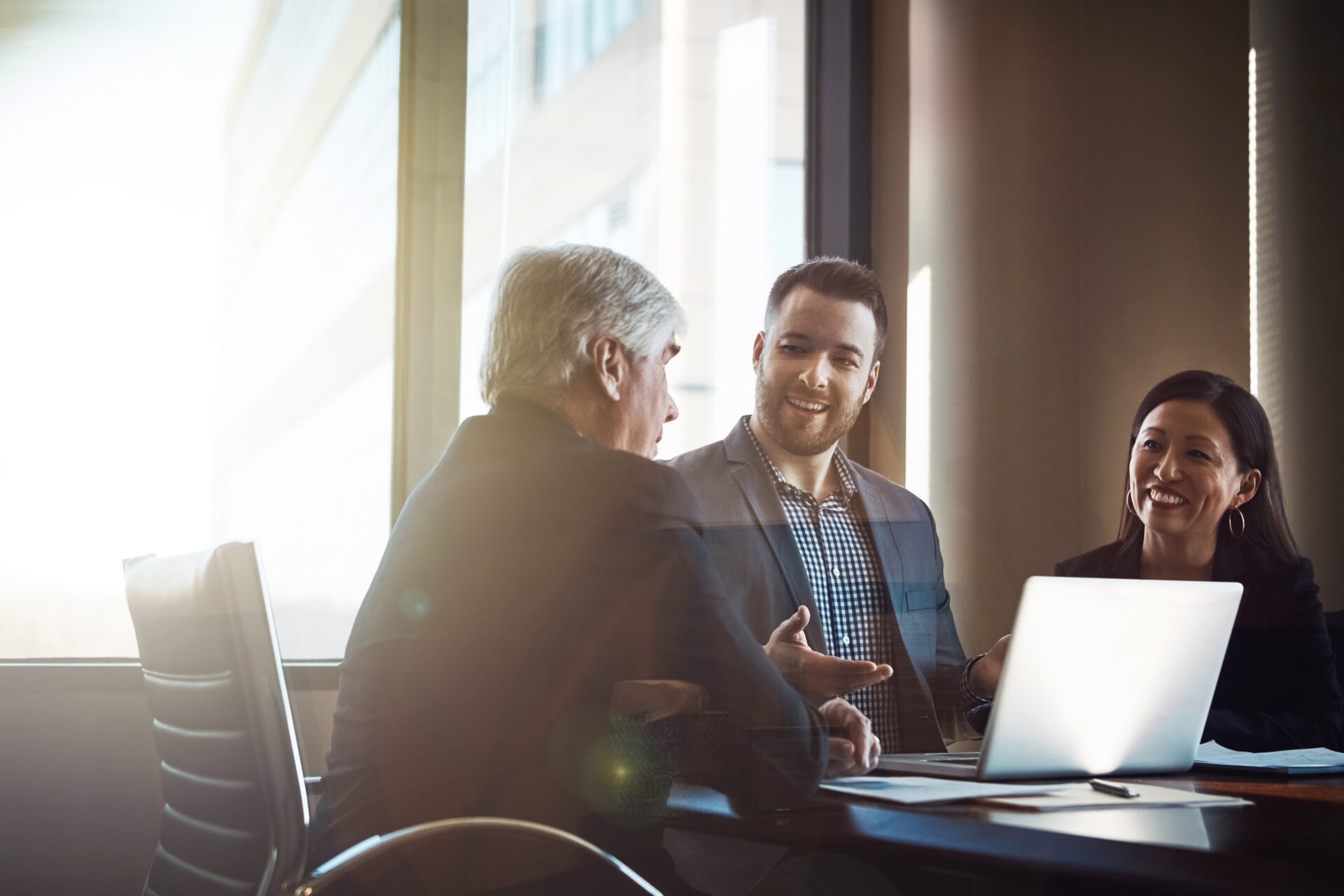 Three businesspeople sit at a table in a bright meeting room, talking with an open laptop and sunlight coming through large windows.
