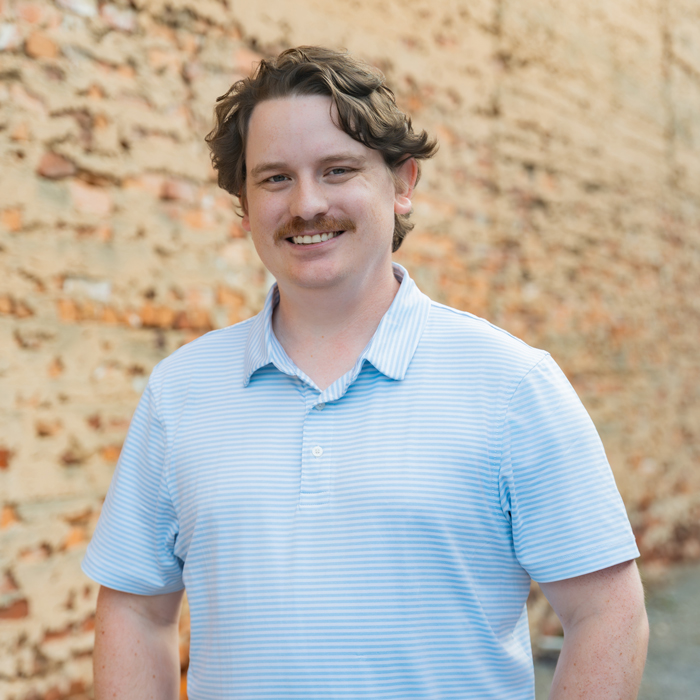 A man with wavy brown hair and a mustache smiles at the camera, wearing a light blue collared shirt before a brick wall.