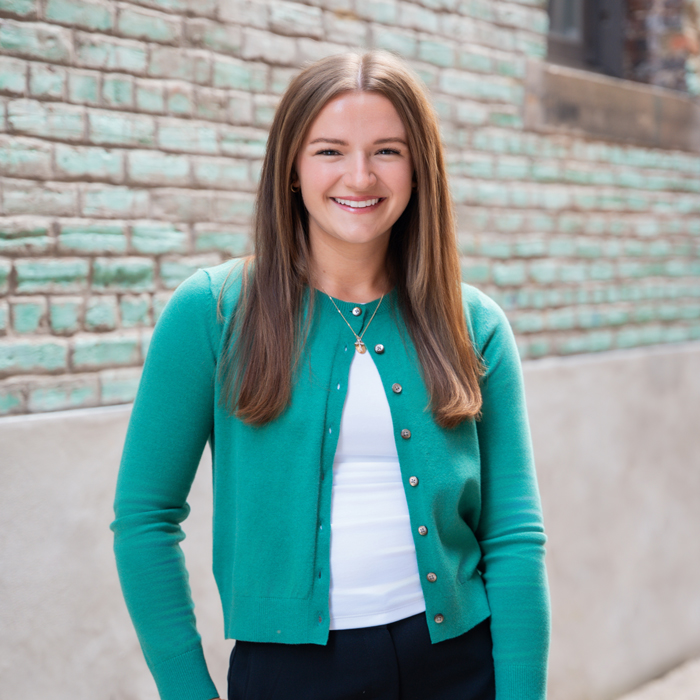 A woman with straight brown hair in a green cardigan, white top, and dark pants smiles outside in front of a brick wall.