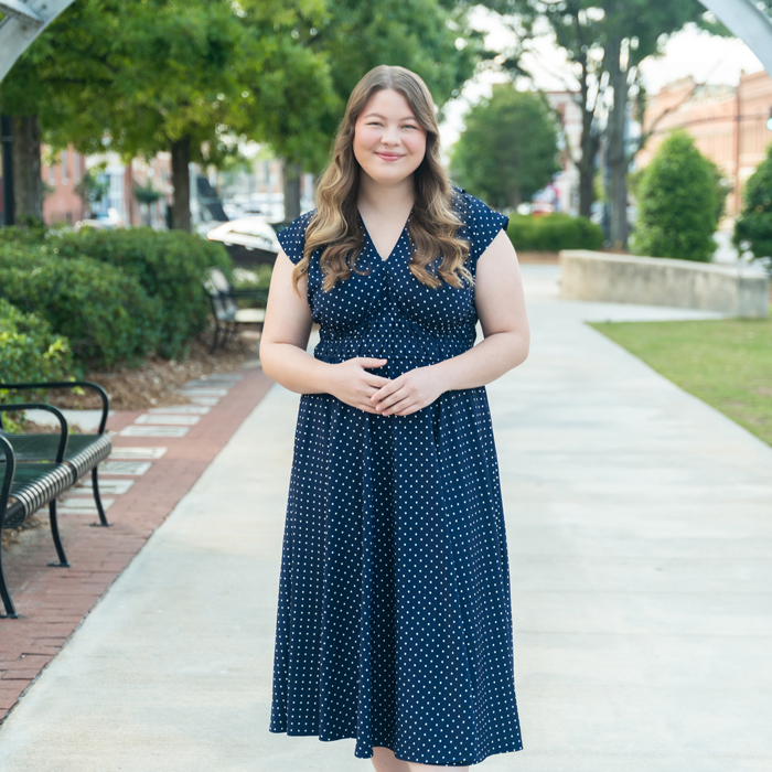 A woman with long wavy hair in a navy polka dot dress smiles on a park sidewalk, trees and benches visible behind her.