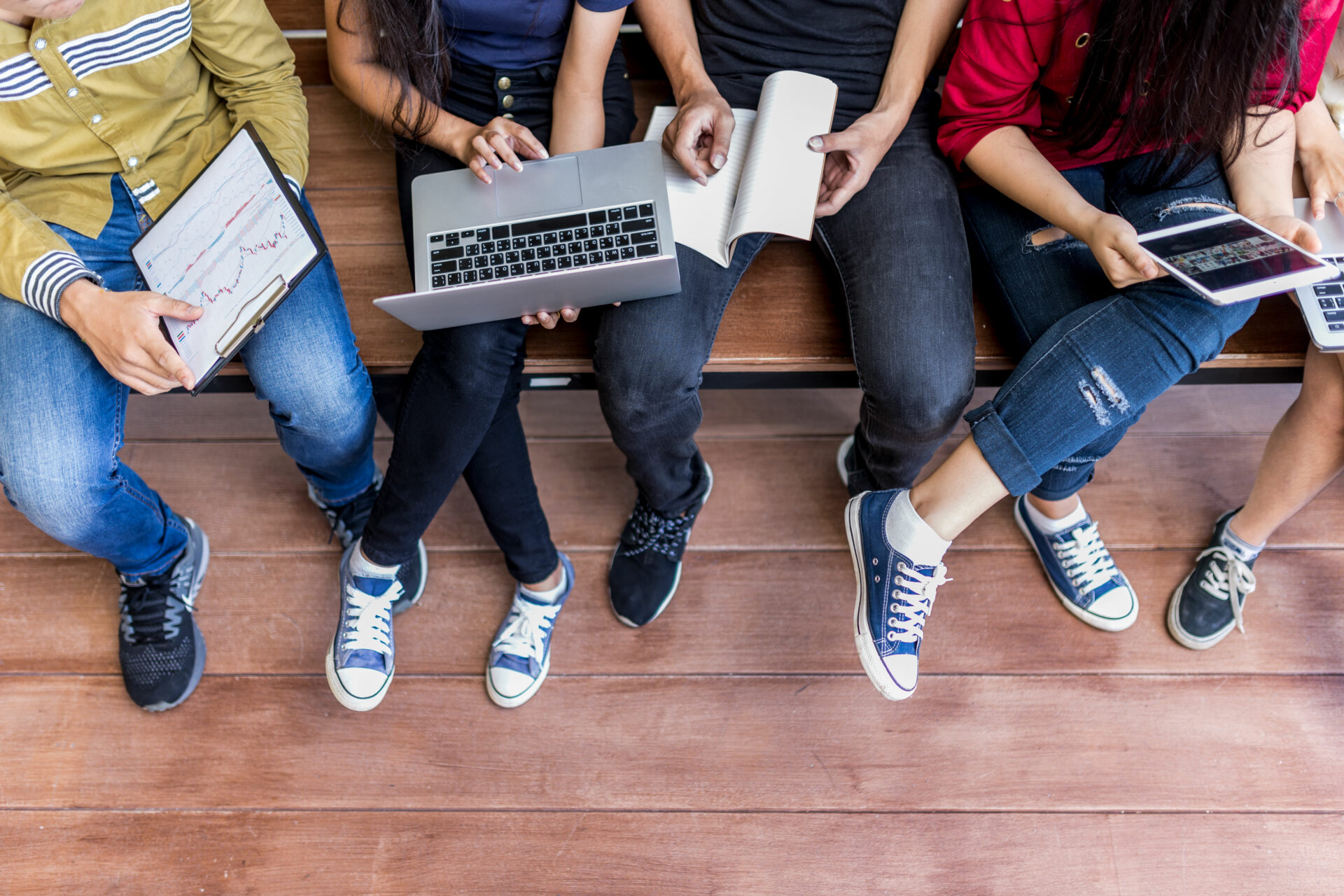 Four people on a bench seen from above using devices and notebooks; one has a tablet with graphs, another types on a laptop, others write or use tablets.