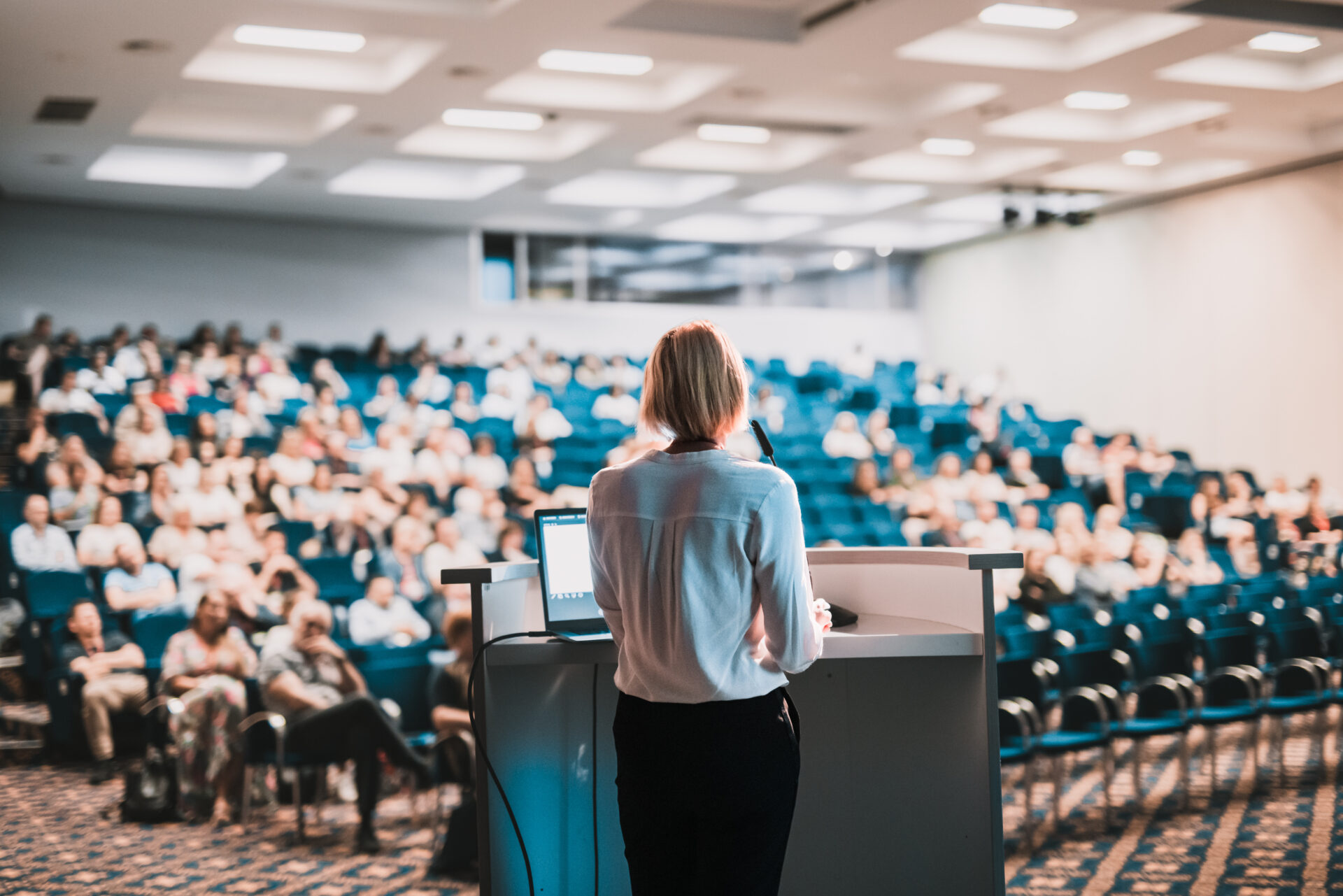 A person presents at a podium to a large audience in a spacious, well-lit conference room with rows of blue chairs.