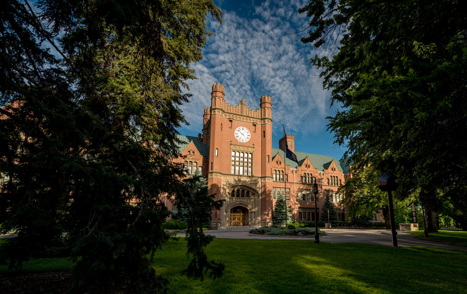 A grand historic brick building features a clock tower, with lush green trees and grass beneath a partly cloudy blue sky.